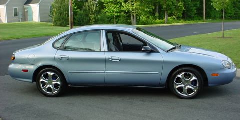 1998 Ford Taurus - A light blue sedan is parked on an asphalt surface with a grassy area and trees in the background.