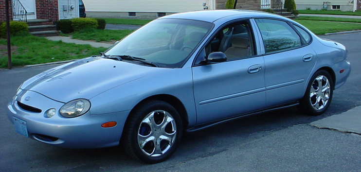 Light blue 1998 Ford Taurus sedan, front 3/4 view, parked on asphalt with a house and lawn in the background