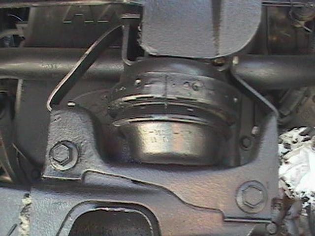 Underside close-up of a round black engine mount in a cast bracket with hex bolts