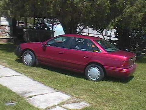 1995 Ford Taurus - A red 1995 Ford Taurus sedan is parked on a grassy area next to a concrete walkway.