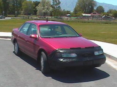 1995 Ford Taurus - A red sedan is parked on asphalt with a black car bra covering the front end.