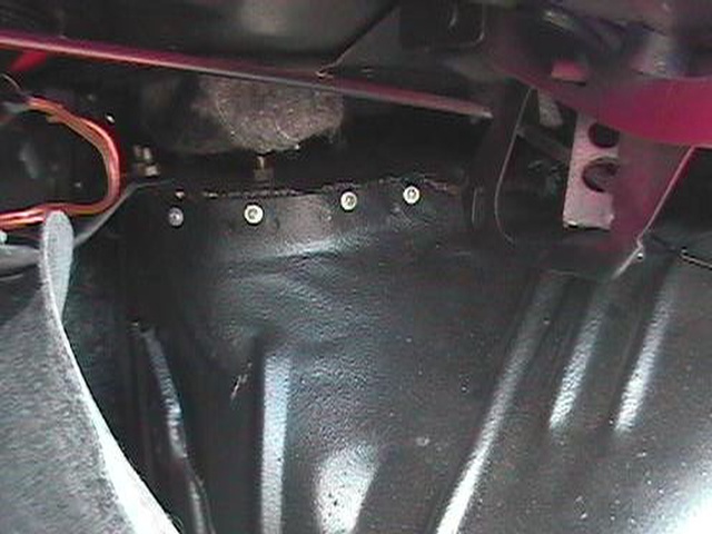 Bare black firewall sheet metal with row of bolts and red wiring inside a car footwell
