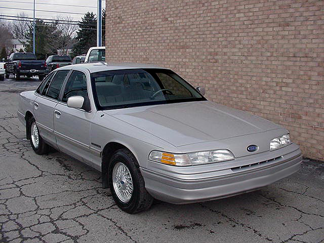 Silver Ford sedan, front 3/4 view, parked by brick building on cracked pavement.
