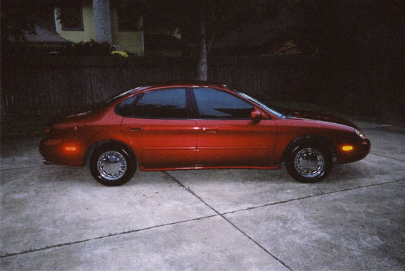 Side profile of red Ford Taurus sedan parked on a driveway by a wood fence