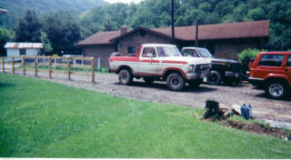 1978 Ford Bronco - Several vehicles are parked in a gravel lot in front of a building with a wooded hillside behind it.