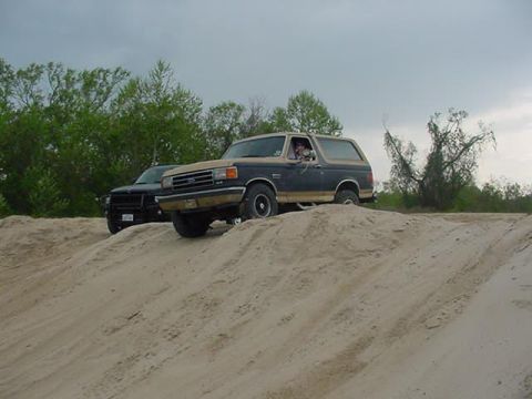 1990 Ford Bronco - A Ford Bronco navigates a sandy terrain, with another vehicle visible in the background under a cloudy sky.
