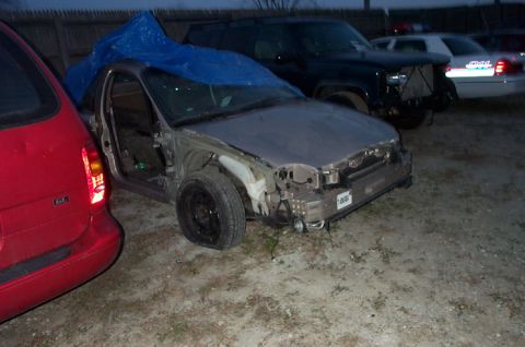 1996 Ford Taurus - A severely damaged car sits in a lot next to a red van, with a dark SUV and sedan in the background.