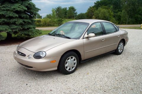 1996 Ford Taurus - A gold sedan is parked on a gravel surface with trees and a cloudy sky in the background.