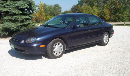 1996 Ford Taurus - A dark blue 1996 Ford Taurus sedan is seen from the front 3/4 angle on a gravel surface with trees in the background.