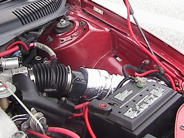 Engine bay with black intake tube, silver heat-wrapped intake, red wiring, and battery in a maroon car