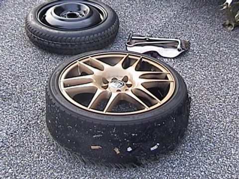 1998 Ford Taurus - A flat tire and a spare tire are shown on a gravel surface, along with a car jack.