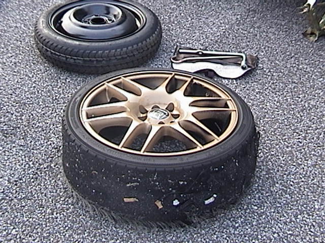 Flat tire with a damaged sidewall next to a spare tire and a jack on the ground