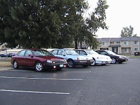 1998 Ford Taurus - A line of sedans are parked in a lot with apartment buildings in the background.