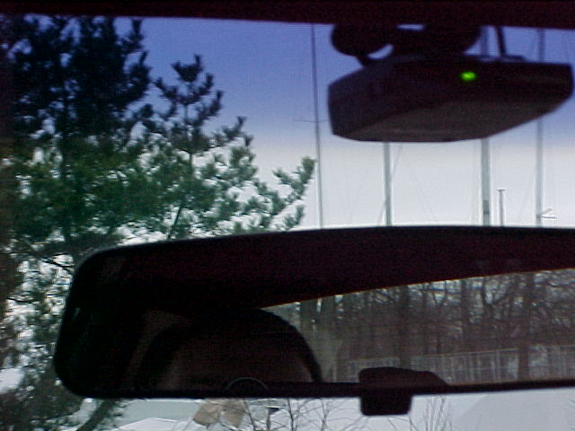 Windshield view from inside a car showing rearview mirror, mounted device with green LED, snowy trees outside