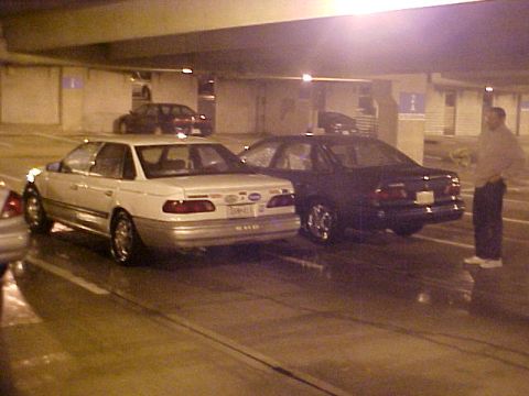 2001 Ford Taurus - Two sedans are parked in a dimly lit indoor parking area. A man stands near the dark car.