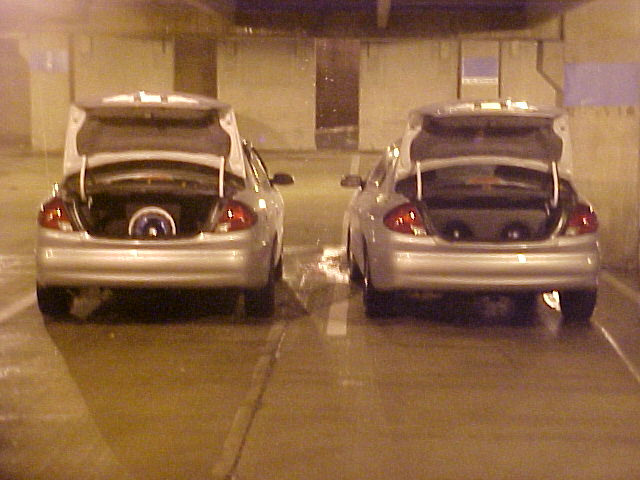 Two silver sedans parked side-by-side with open trunks, showing custom speaker setups inside