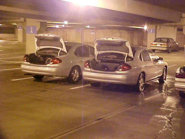 Two silver sedans with trunks open parked side by side in a covered parking garage at night