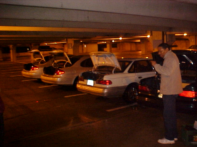 Several sedans parked in a garage, some with trunks open. A man stands near one of the cars