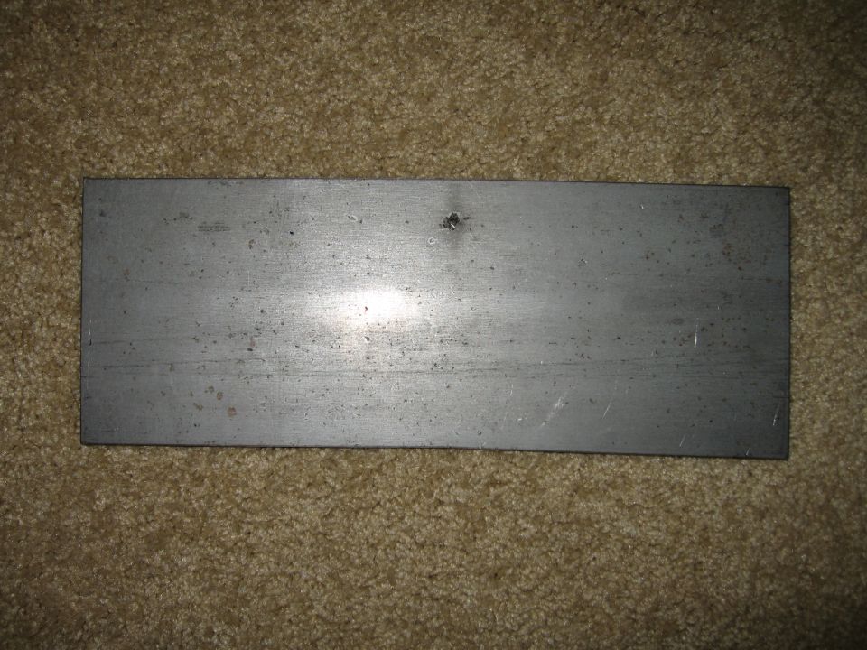 Flat rectangular sheet metal plate with single hole resting on carpet