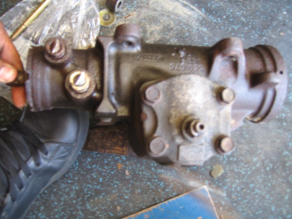 Close-up of a power steering gearbox with part number 9491699 visible. A hand and a black shoe are in the frame