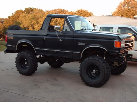 1991 Ford Bronco - Passenger-side profile of a dark blue-gray lifted Ford Bronco with its hardtop removed, running large mud-terrain tires on black steel whee…