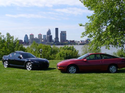 1991 Ford Probe - A red first-generation Ford Probe hatchback coupe on stock silver alloys parked on grass beside a black Mazda RX-8, with a river and city s…