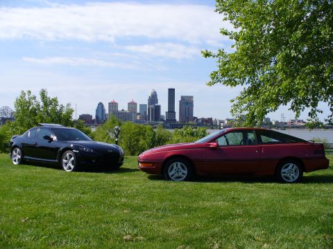 1991 Ford Probe - A red Ford Probe hatchback coupe on silver five-spoke alloys parked on riverfront grass beside a black Mazda RX-8, with a city skyline and…