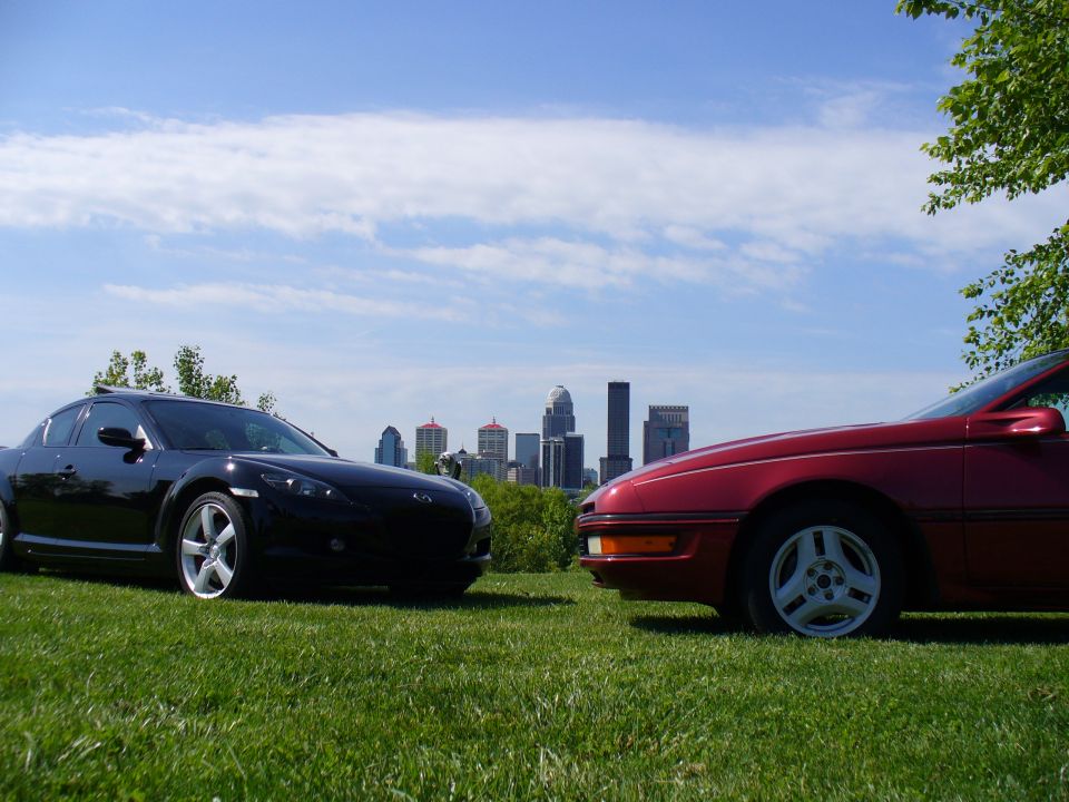 Black Mazda RX-8 and maroon Ford Probe coupes on grass with a city skyline behind