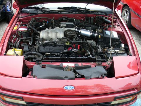 1991 Ford Probe - Engine bay of a red Ford, viewed from the front with the hood open, showing a transversely-mounted 3.0L V6 engine with a black aftermarket…