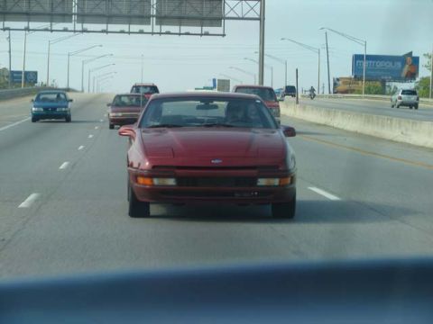 1991 Ford Probe - Rolling shot taken through a windshield of a maroon first-generation Ford Probe traveling ahead on a multi-lane highway, seen head-on with…