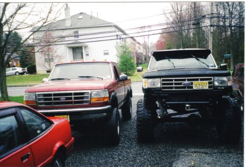 1988 Ford Bronco - Two lifted trucks, a red Ford and a black Ford F-Series, are parked side-by-side.