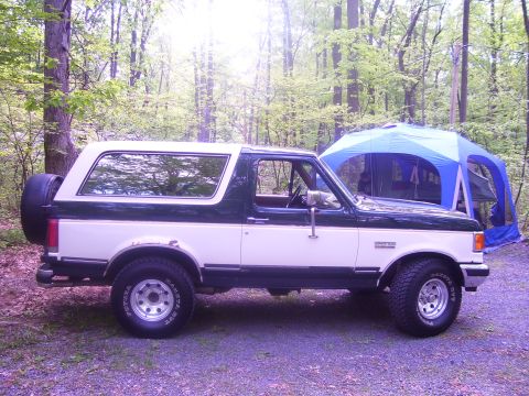 1991 Ford Bronco - Side view of a two-tone 1991 Ford Bronco parked in a forest campsite.
