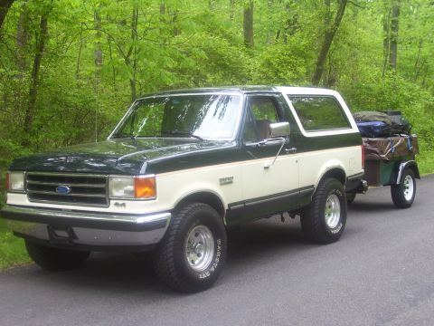 1991 Ford Bronco - A two-tone green and white 1991 Ford Bronco is parked on a paved surface, towing a trailer filled with bags.