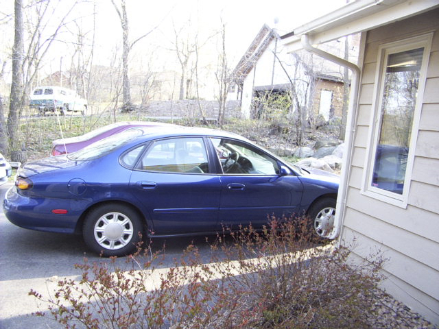 Blue Ford Taurus sedan parked beside a tan house with bare winter trees