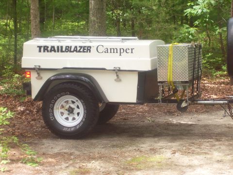1991 Ford Bronco - Side view of a small cream and black single-axle Trailblazer Camper cargo trailer parked on a wooded gravel site, with a hinged clamshell l…