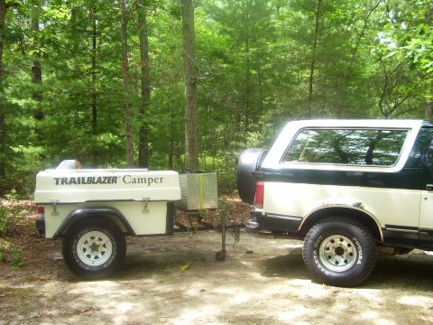 1991 Ford Bronco - A green-over-white two-tone full-size Ford Bronco on Dakota A/T tires and white wagon wheels parked at a wooded campsite, with a rear-mount…