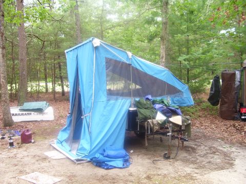 1991 Ford Bronco - A partially set-up blue pop-up tent camper trailer at a wooded campsite, with green and blue gear bags loaded on the trailer bed and its A-…
