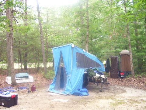 1991 Ford Bronco - A small tent-trailer camper set up at a wooded campsite, its light blue canvas tent erected over the trailer frame with the tongue and jack…