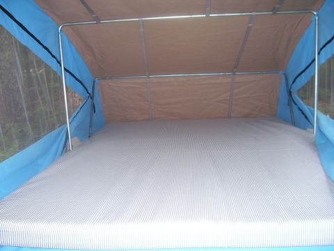 1991 Ford Bronco - Interior of a pop-up camper bunk end, showing a bare striped mattress under a tan canvas canopy supported by metal tube framing, with blue…