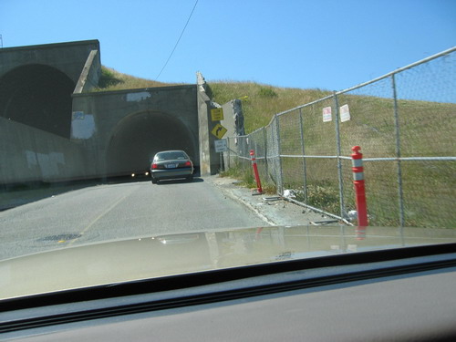 Dark sedan seen from behind exiting a tunnel, with a chain-link fence and orange bollards to the right