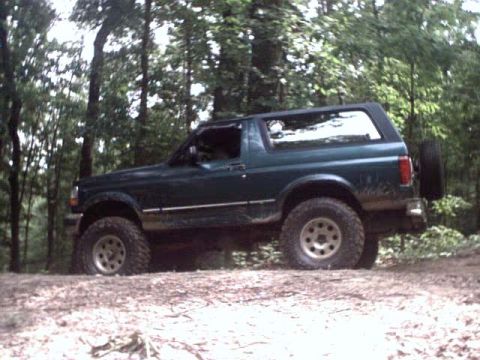 1996 Ford Bronco - A lifted, dark green Ford Bronco with off-road tires navigates a dirt path through a wooded area.