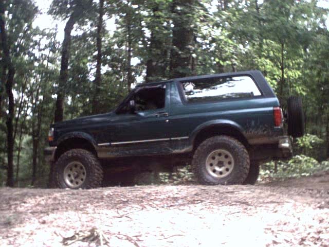 Dark green Ford Bronco, lifted, off-road tires, spare tire on rear door, driving on dirt road in woods