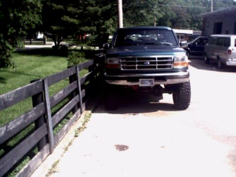 1996 Ford Bronco - A green Ford F-Series truck is parked in front of a building, with a wooden fence to its left.
