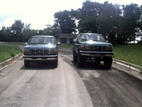1996 Ford Bronco - Two Ford Broncos are parked on a paved road with trees and a house in the background.