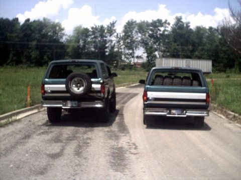 1996 Ford Bronco - Two 1996 Ford Broncos are seen from the rear, driving on a dirt road. The Bronco on the left has a spare tire mounted on the tailgate.