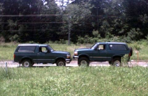 1996 Ford Bronco - Two dark green Ford Broncos are parked next to each other on a grassy roadside.