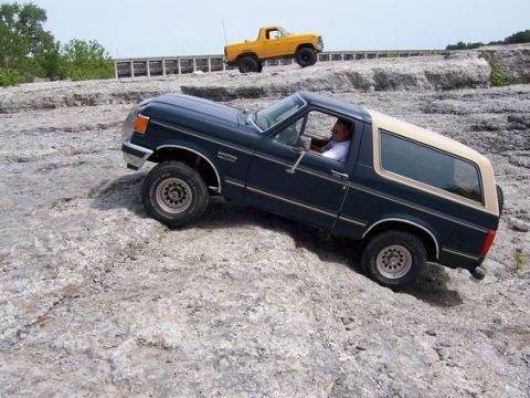 1991 Ford Bronco - A Ford Bronco navigates a rocky terrain, with another Ford truck visible in the background.