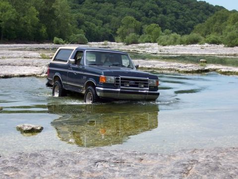 1991 Ford Bronco - A dark blue 1991 Ford Bronco navigates a rocky riverbed on a sunny day.
