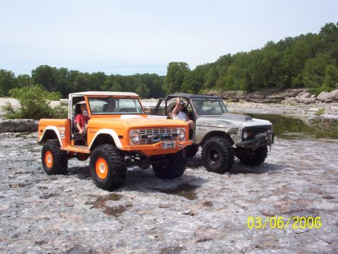 1991 Ford Bronco - Two Ford Broncos are parked on a rocky riverbed under a cloudy sky.