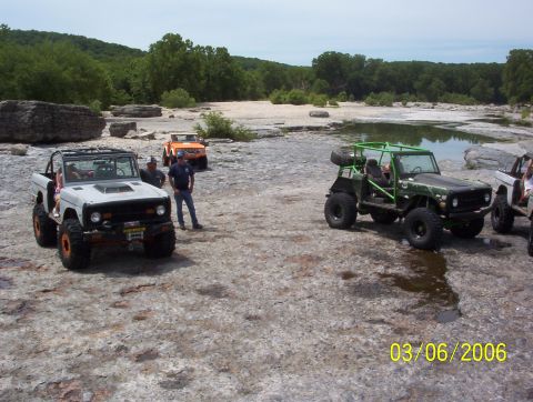 1991 Ford Bronco - Several modified off-road vehicles are parked on a rocky landscape with trees and water in the background.